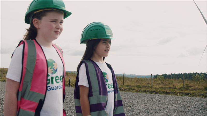 Two young girls with green hard hats, wearing green guardians t shirt standing at teiges wind farm grounds